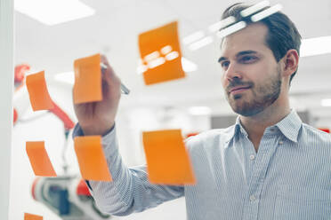 Smiling young male engineer writing on adhesive notes sticked at glass wall - DIGF14585