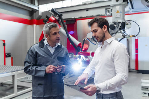 Junger männlicher Ingenieur, der ein digitales Tablet benutzt, während er sich mit einem reifen Mann in einer Fabrik unterhält - DIGF14607