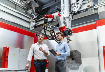 Mature automation engineer explaining robotic to young man with digital tablet in factory - DIGF14646