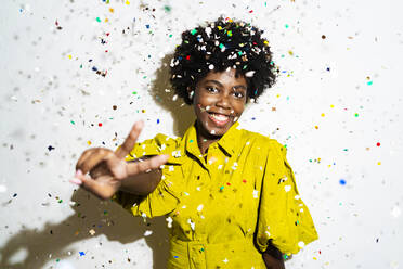 Cheerful curly hair woman showing peace sign while standing amidst confetti against white background - GIOF11397