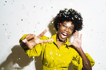 Confetti falling on woman gesturing peace sign while standing white background - GIOF11398