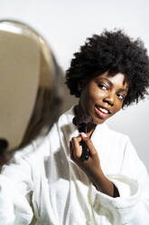 Young woman with hand mirror doing make-up while standing against white background - GIOF11401
