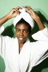 Lächelnde Frau im Bademantel, die ein Handtuch bindet, während sie vor einem grünen Hintergrund steht - GIOF11407