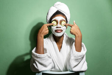 Junge Frau im Bademantel, die ihre Augen mit einer Gurke bedeckt, während sie vor einem grünen Hintergrund steht - GIOF11419