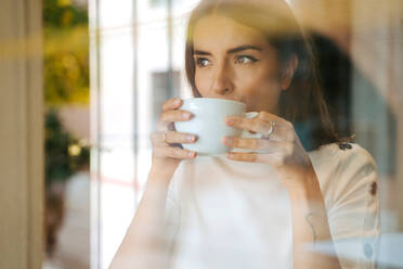 Blick durch das Fenster auf eine ruhige junge Frau, die frischen aromatischen Kaffee aus einer weißen Tasse trinkt und wegschaut, während sie den Morgen zu Hause verbringt - ADSF20989