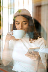 Blick durch eine Glasscheibe auf eine fröhliche junge Frau, die auf ihrem Handy surft, während sie mit einer Tasse Kaffee am Fenster steht und die freie Zeit zu Hause genießt - ADSF20990
