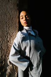 Serene young ethnic female in stylish gray coat leaning back against aged stone wall and enjoying sunlight - ADSF20991