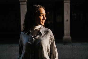 Moderne ethnische Frau in modischem Mantel und mit Tasche, die in der Fußgängerzone geht und in die Kamera schaut - ADSF20995