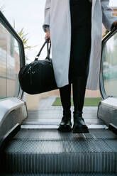 Gesichtslose elegante Geschäftsfrau in Mantel und mit Tasche, die eine Rolltreppe im Stadtzentrum betritt - ADSF20998