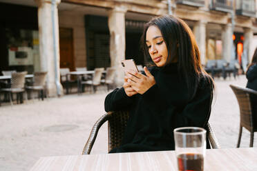 Relaxed ethnic female in black clothes having drink at cafe table on street and texting via smartphone - ADSF21000