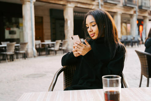 Entspannte ethnische Frau in schwarzer Kleidung, die in einem Straßencafé einen Drink zu sich nimmt und über ihr Smartphone eine SMS schreibt - ADSF21000