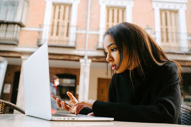 Seitenansicht einer ethnischen Frau im schwarzen Pullover, die am Tisch eines Straßencafés an einem Netbook arbeitet und tippt - ADSF21004