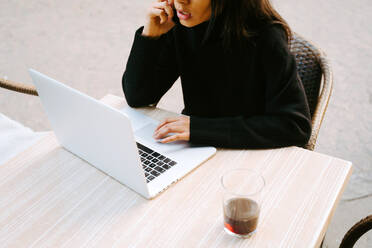 Ethnische Frau in schwarzem Pullover arbeitet an einem Netbook am Tisch eines Straßencafés und tippt - ADSF21005