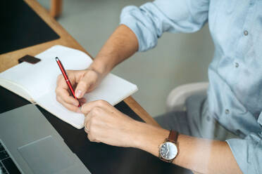 Unbekannter männlicher Unternehmer sitzt am Tisch und schreibt in ein Notizbuch, während er ein Geschäftsprojekt plant - ADSF21029