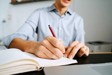 Unbekannter männlicher Unternehmer sitzt am Tisch und schreibt in ein Notizbuch, während er ein Geschäftsprojekt plant - ADSF21030