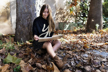 Full body of peaceful stylish female teenager with long hair in casual outfit sitting near tree amidst fallen trees and reading interesting book in autumn park - ADSF21036