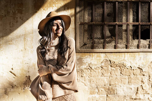 Fashionable young smiling ethnic female with long dark hair in stylish warm winter outfit and hat standing in autumn park near aged house with shabby walls looking away - ADSF21040