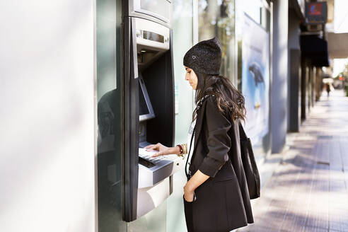 Seitenansicht einer jungen, trendigen, ethnischen Dame in Freizeitkleidung und Hut, die den PIN-Code eingibt, während sie einen Geldautomaten benutzt, der an einem sonnigen Tag auf einer Straße in der Stadt steht - ADSF21042