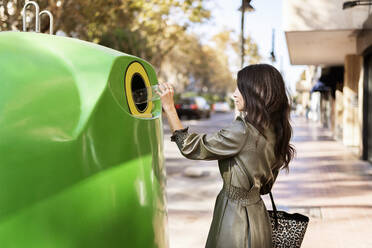 Seitenansicht von jungen stilvollen ethnischen Frau in Lederkleid werfen Plastikflasche in grünen Recycling-Behälter auf der Straße platziert Stadt - ADSF21043