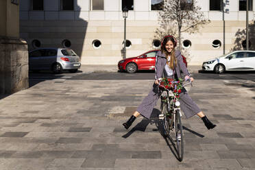 Ganzer Körper einer glücklichen jungen Frau in stilvoller Freizeitkleidung und mit Kopfhörern, die Spaß haben, während sie auf einem mit Blumen geschmückten Fahrrad auf einem gepflasterten Platz in der Stadt fährt - ADSF21051