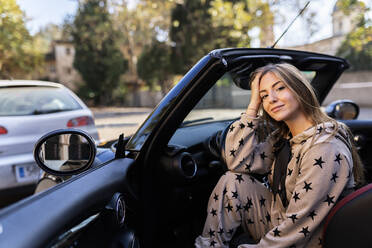 Smiling millennial female in stylish apparel sitting on driver seat of luxurious convertible automobile parked on city street and looking at camera - ADSF21054