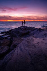 Rückansicht der Silhouette eines anonymen Touristenpaares, das auf einem Felsen steht und den malerischen Sonnenuntergang über dem Meer im Sommer in Cádiz beobachtet - ADSF21073