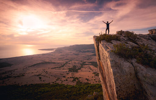 Rückansicht der Silhouette eines anonymen Touristen, der auf einem felsigen Hügel steht und in die Ferne zeigt, während er den atemberaubenden Sonnenuntergang über dem Meer in Cádiz bewundert - ADSF21076