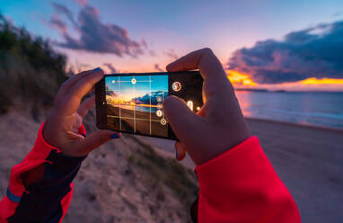 Rückenansicht einer nicht erkennbaren weiblichen Reisenden mit langen dunklen Haaren in Freizeitkleidung, die mit ihrem Smartphone während ihres Urlaubs in Kantabrien einen wunderschönen Sonnenuntergang über dem Meer mit Sandstrand fotografiert - ADSF21082