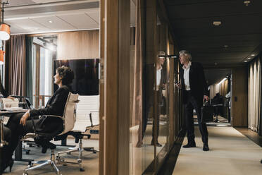 Curious businessman looking through glass wall at colleagues in board room during meeting - MASF22011