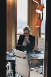 Geschäftsfrau, die einen Laptop benutzt, während sie am Konferenztisch im Sitzungssaal sitzt, gesehen durch eine Glaswand - MASF22041