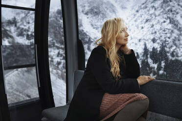 Contemplated blond woman looking through window while sitting in overhead cable car - AZF00200