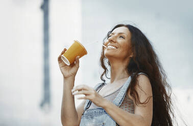 Fröhliche Frau trinkt Kaffee aus einem Einweg-Kaffeebecher in der Stadt - AZF00205