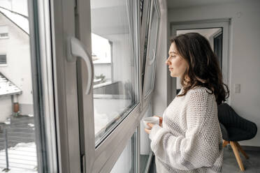 Frau steht mit Kaffeetasse und schaut durch das Fenster zu Hause - OGF00909