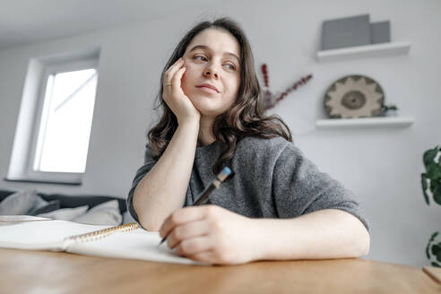 Nachdenkliche Frau sitzt am Tisch und schreibt in ein Notizbuch im Wohnzimmer - OGF00913