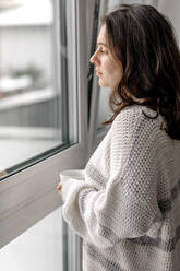 Frau im Pullover mit Kaffeetasse stehend, während sie durch das Fenster zu Hause schaut - OGF00914