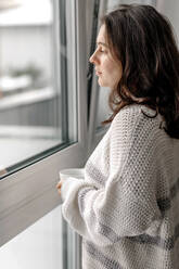 Woman in sweater standing with coffee cup while looking through window at home - OGF00914