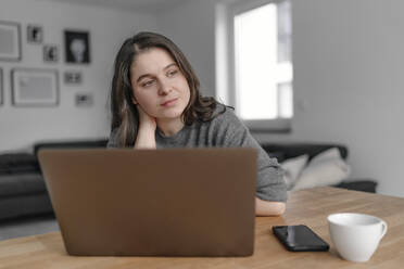 Nachdenkliche weibliche Berufstätige, die mit ihrem Laptop zu Hause sitzt und wegschaut - OGF00917