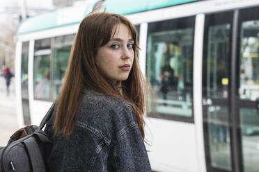 Junge Frau mit braunen Haaren steht an einer Straßenbahnhaltestelle vor einem Zug - PNAF00839