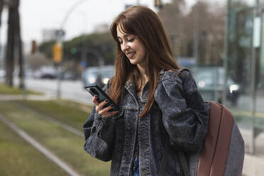 Lächelnde Frau, die ein Smartphone benutzt, während sie an einer Straßenbahnhaltestelle steht - PNAF00842