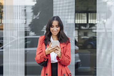 Smiling woman in red trench coat using smart phone while standing against glass wall - JRVF00315