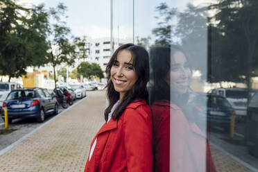 Smiling young woman in red trench coat leaning on glass window - JRVF00316