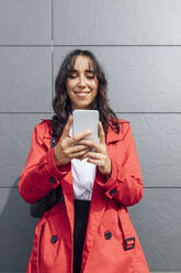 Lächelnde junge Geschäftsfrau, die telefoniert, während sie vor einer grauen Wand steht - JRVF00317