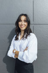 Smiling female entrepreneur with arms crossed standing against gray wall - JRVF00323