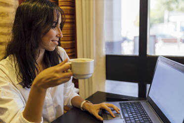 Junge Geschäftsfrau arbeitet an einem Laptop und hält eine Tasse Kaffee in einem Cafe - JRVF00335