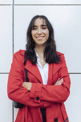 Lächelnde junge Frau in rotem Trenchcoat steht mit verschränkten Armen vor einer weißen Wand - JRVF00336