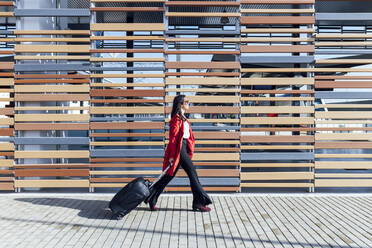 Businesswoman walking on footpath with suitcase by wooden wall during sunny day - JRVF00340