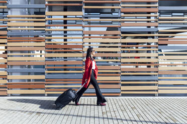 Geschäftsfrau, die auf einem Fußweg mit einem Koffer an einer Holzwand bei Sonnenschein spazieren geht - JRVF00340