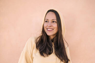 Beautiful smiling woman wearing hoodie while looking away against wall - GRCF00687