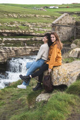 Glückliche Freundinnen sitzen auf einem Felsen am Wasserfall - JAQF00328