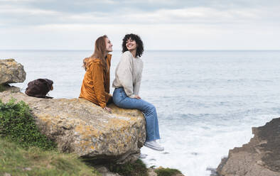 Female friends smiling looking at each other while sitting on cliff by sea - JAQF00329